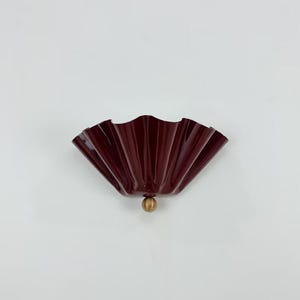 May include: A burgundy wall sconce with a pleated, fan-shaped design. It features a brass-coloured spherical accent at the base. The fixture is mounted on a white wall, providing a striking contrast.