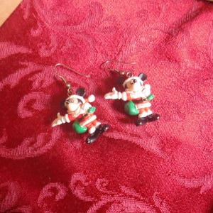 Peut inclure: Une paire de boucles d'oreilles pendantes repr&eacute;sentant Mickey Mouse v&ecirc;tu d'un costume de P&egrave;re No&euml;l avec un manteau rouge, une bordure blanche, une ceinture noire et des bottes noires, portant un sac vert. Les boucles ont des crochets en m&eacute;tal argent&eacute;.