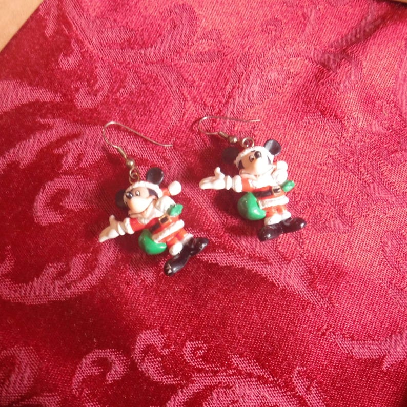 Peut inclure: Une paire de boucles d'oreilles pendantes repr&eacute;sentant Mickey Mouse v&ecirc;tu d'un costume de P&egrave;re No&euml;l avec un manteau rouge, une bordure blanche, une ceinture noire et des bottes noires, portant un sac vert. Les boucles ont des crochets en m&eacute;tal argent&eacute;.