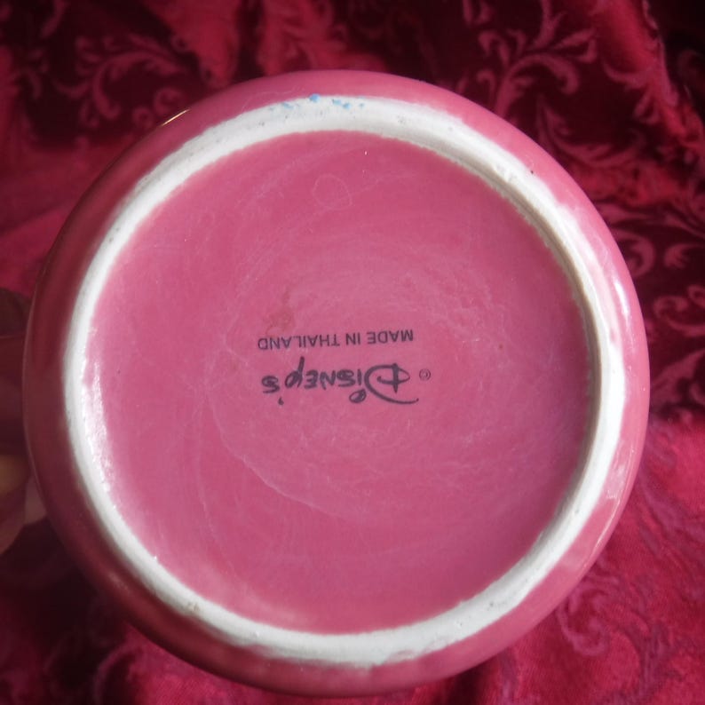 May include: The circular base of a pink ceramic mug featuring a white rim. Printed in black text on the bottom is the Disney logo followed by the words MADE IN THAILAND. The surface is smooth with a glossy finish and a small blue mark near the edge.