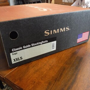 May include: A rectangular cardboard box for Simms Classic Guide Stockingfoots in sage color, size XXLS. The box is black with a tan top, featuring the Simms logo, a small American flag graphic, a circular cutout, and a product label with a barcode.