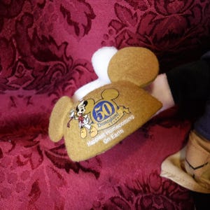 May include: A tan felt ear hat featuring a graphic of Mickey Mouse and the text 50 Disneyland Resort Happiest Homecoming On Earth. The hat has two round ears and a white trim detail on the top edge, showcasing a commemorative design.