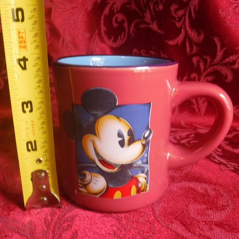 May include: A pink ceramic mug featuring a graphic of Mickey Mouse on the front. The mug has a light blue interior and a dark purple rim. A yellow measuring tape is positioned next to the mug, showing a height of approximately 4 inches.