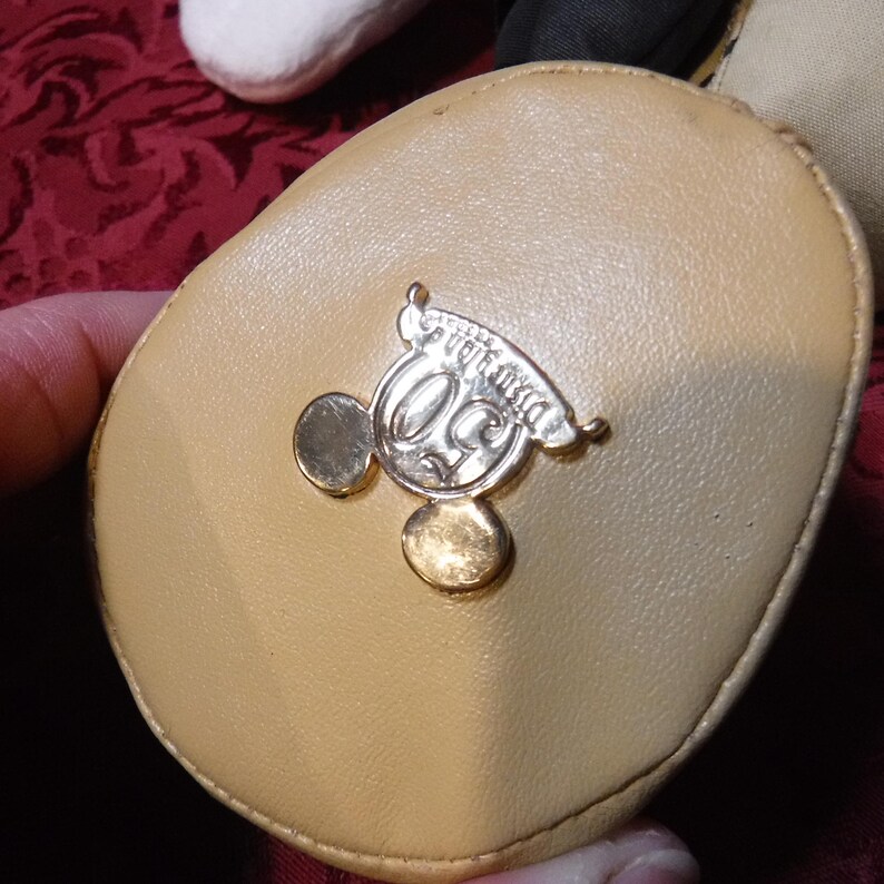 May include: A tan, oval-shaped fabric patch featuring a metallic gold-tone emblem. The emblem displays a stylized number 50 with two circular ears resembling a mouse silhouette, and a small banner above the number with the text Walt Disney World.