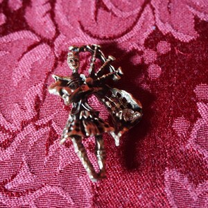 May include: A vintage-style brooch in the shape of a bagpiper playing the bagpipes. The brooch is made of a gold-colored metal and features intricate details of the bagpiper's clothing and the instrument. The brooch is set against a red patterned background.