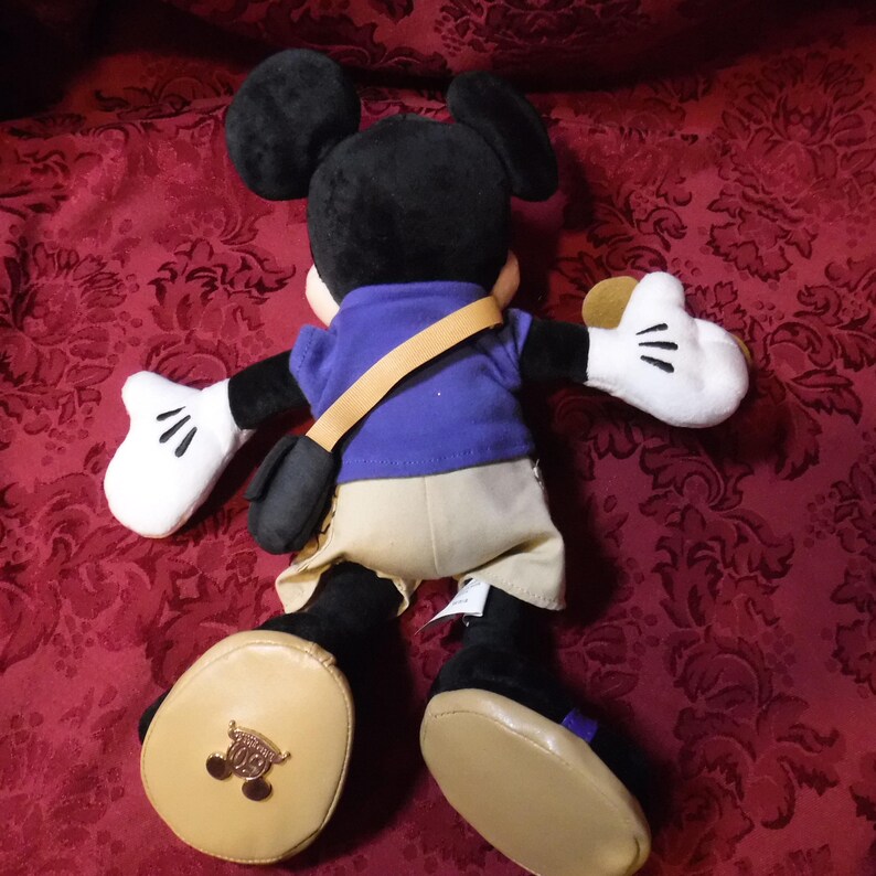 May include: A plush Mickey Mouse toy wearing a purple shirt, tan shorts, and a tan crossbody bag. The character has black ears, white gloves, and tan shoes. A gold-colored emblem featuring the Mickey Mouse silhouette is attached to the left shoe.