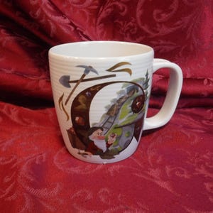 May include: A white ceramic mug with a ribbed texture featuring a graphic of a bearded character with a red hat holding a gem. The design includes a large letter G, a mine cart on tracks, pickaxes, and various colorful gemstones against a landscape.