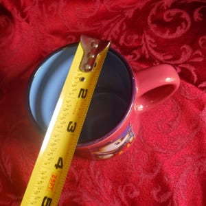 May include: A pink ceramic mug with a light blue interior and a cartoon character graphic on the side. A yellow measuring tape is placed across the top opening, showing a diameter of approximately 3.5 inches for the mug's rim.