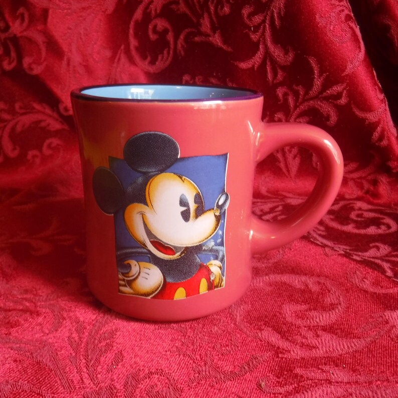 May include: A coral-colored ceramic mug featuring a graphic of Mickey Mouse on the front. The mug has a rounded handle, a smooth exterior finish, and a light blue interior rim. The character graphic shows Mickey Mouse with a smiling expression.
