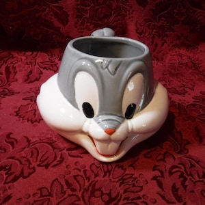 May include: A ceramic mug shaped like Bugs Bunny's head. The mug features a gray top with large white eyes, black pupils, and a red nose. The bottom half is white, with Bugs' signature buck teeth. A small gray handle is on the top.