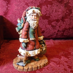 May include: A detailed figurine of Santa Claus, wearing a red coat with white trim and a matching hat. He carries a basket of fruit and a sack with a snowman. A small reindeer on a wheeled cart stands at his feet. The base is a cobblestone design.