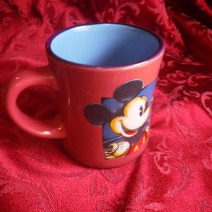 May include: A ceramic mug with a coral-red exterior and a light blue interior. The mug features a graphic illustration of Mickey Mouse on the side, depicted with his signature black ears, white face, and red shorts, set against a dark blue square.