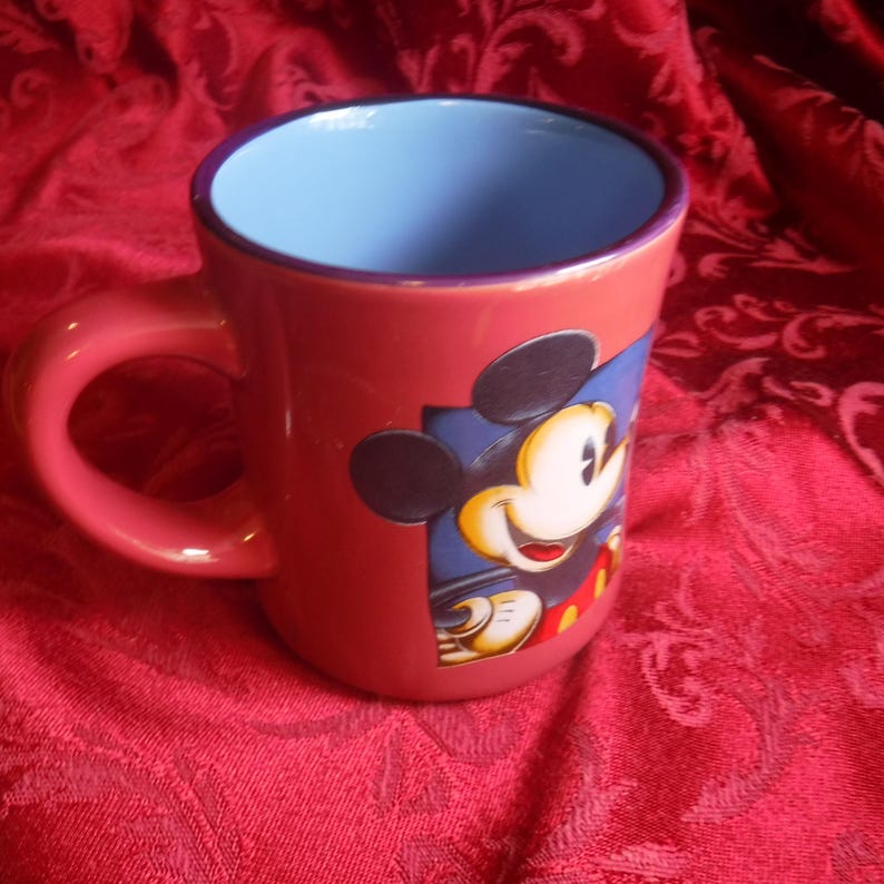 May include: A ceramic mug with a coral-red exterior and a light blue interior. The mug features a graphic illustration of Mickey Mouse on the side, depicted with his signature black ears, white face, and red shorts, set against a dark blue square.