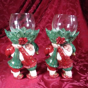 May include: A pair of ceramic candle holders featuring a whimsical design of a gnome holding a holly-adorned glass votive. The gnomes are dressed in red and green with gold accents.