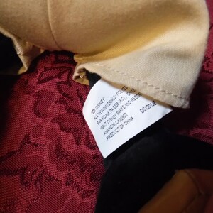 May include: A white fabric tag attached to a plush toy with black and tan material. The tag features black text reading Disney, All New Materials: Polyester Fiber, Eva Foam, PA 8336 (RC), OH18442, Walt Disney Parks and Resorts, Anaheim, CA 92803, Product of China, DS1201JE.