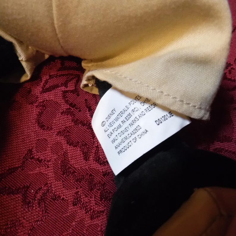 May include: A white fabric tag attached to a plush toy with black and tan material. The tag features black text reading Disney, All New Materials: Polyester Fiber, Eva Foam, PA 8336 (RC), OH18442, Walt Disney Parks and Resorts, Anaheim, CA 92803, Product of China, DS1201JE.