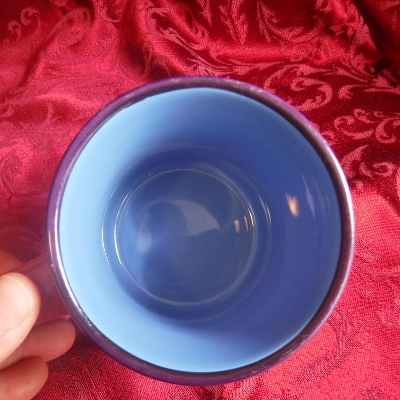 May include: A circular mug viewed from directly above, featuring a smooth, light blue interior and a deep purple exterior rim. The mug has a simple, cylindrical shape with a solid color finish on the inside and a dark, reflective outer edge.
