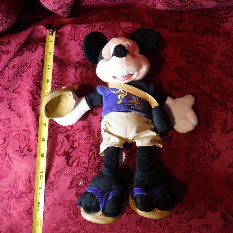 May include: A Mickey Mouse plush toy wearing a purple shirt with a 50th anniversary graphic, tan shorts, and sandals. A yellow measuring tape placed next to the plush shows a height of approximately 11 inches from the top of the ears to the feet.