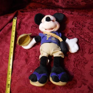 May include: A plush Mickey Mouse toy wearing a purple shirt with the text 'Happiest Homecoming On Earth' and a 50th anniversary logo, tan shorts, and purple flip-flops, holding a tan hat, next to a yellow measuring tape showing 11 inches.