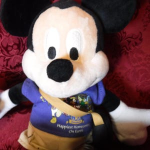 May include: A plush Mickey Mouse toy wearing a purple t-shirt with a graphic print and the text Disneyland 50th Happiest Homecoming On Earth, tan shorts, and a tan crossbody bag. The character has black ears, a white face, and a black nose.
