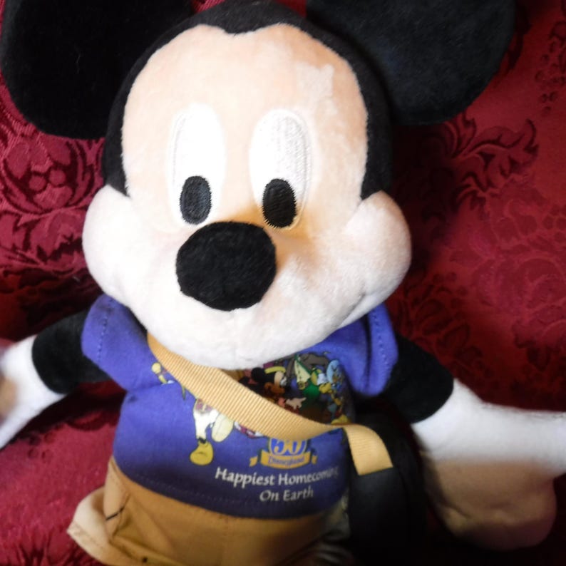 May include: A plush Mickey Mouse toy wearing a purple t-shirt with a graphic print and the text Disneyland 50th Happiest Homecoming On Earth, tan shorts, and a tan crossbody bag. The character has black ears, a white face, and a black nose.