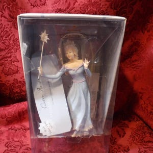 May include: A Hallmark Keepsake ornament of Queen Aurora from Sleeping Beauty, in a clear plastic box. The figurine is wearing a light blue gown with a pink trim and holding a star-topped wand. The text on the tag reads "Queen Aurora".
