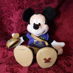May include: A plush Mickey Mouse doll wearing a purple shirt with the text Happiest Homecoming On Earth and a 50th anniversary logo. The doll has tan shoes, one of which features a gold 50th anniversary emblem, and holds a matching tan hat.