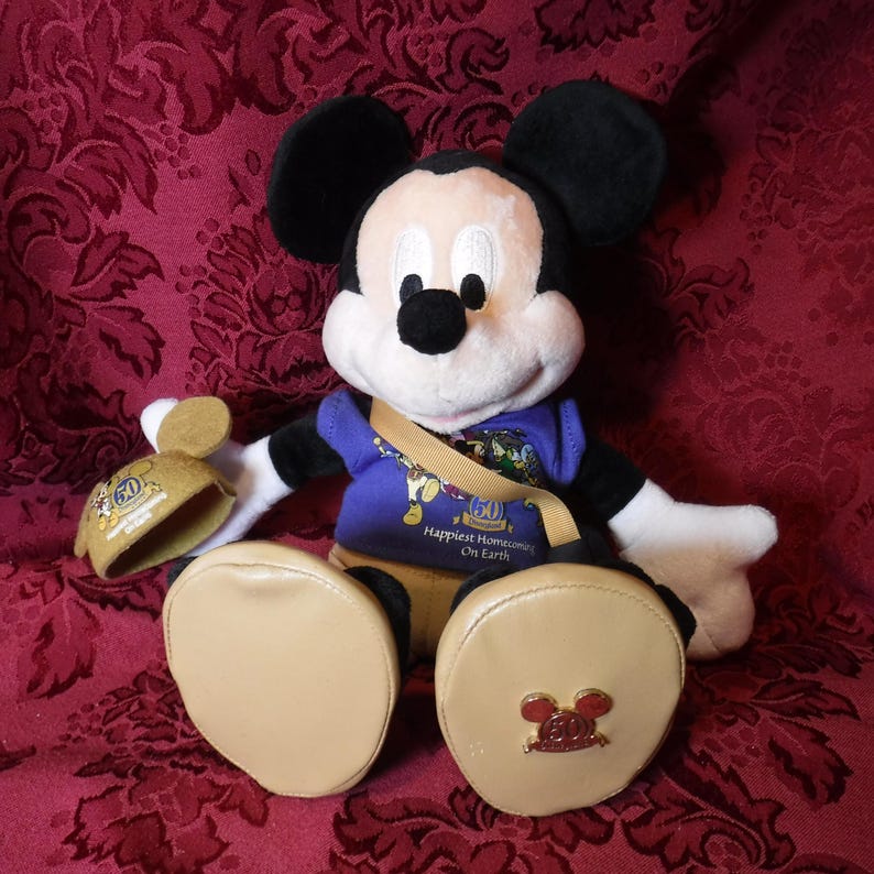 May include: A plush Mickey Mouse doll wearing a purple shirt with the text Happiest Homecoming On Earth and a 50th anniversary logo. The doll has tan shoes, one of which features a gold 50th anniversary emblem, and holds a matching tan hat.