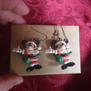 Peut inclure: Une paire de boucles d'oreilles pendantes repr&eacute;sentant Mickey Mouse v&ecirc;tu d'un costume de P&egrave;re No&euml;l rouge et blanc avec un sac vert. Les figurines sont fix&eacute;es &agrave; des crochets en m&eacute;tal argent&eacute;, montrant le personnage avec des oreilles et des chaussures noires.