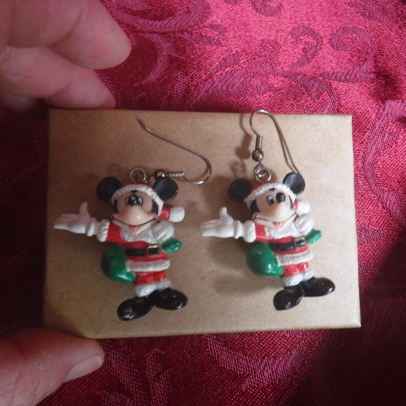 Peut inclure: Une paire de boucles d'oreilles pendantes repr&eacute;sentant Mickey Mouse v&ecirc;tu d'un costume de P&egrave;re No&euml;l rouge et blanc avec un sac vert. Les figurines sont fix&eacute;es &agrave; des crochets en m&eacute;tal argent&eacute;, montrant le personnage avec des oreilles et des chaussures noires.