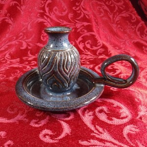 May include: A blue and brown speckled ceramic candle holder with a circular base, a central socket for a candle, and a curved handle. The exterior of the central socket features a raised, vertical flame-like pattern in a dark brown glaze.