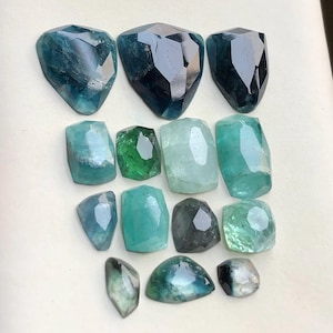 Peut inclure: Une sélection de pierres précieuses facettées dans des tons de bleu, de turquoise et de vert. Les pierres sont présentées sur une surface claire, mettant en valeur leurs facettes polies et leur clarté. Différentes formes, comme des coupes en bouclier et rectangulaires. Mesures en cm.