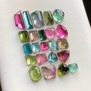 Puede incluir: Una colección de gemas facetadas en varios colores, incluyendo rosa, azul, verde y amarillo. Las gemas están talladas en diferentes formas, como rectángulos y formas irregulares. Las gemas se exhiben sobre una superficie blanca.