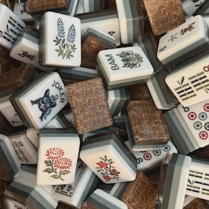 May include: A collection of rectangular Mahjong tiles featuring white faces with various illustrations including a blue lupine flower, a green dragon, a French bulldog labeled JOKER, red chrysanthemums, and bamboo patterns, with brown woven-textured backs.