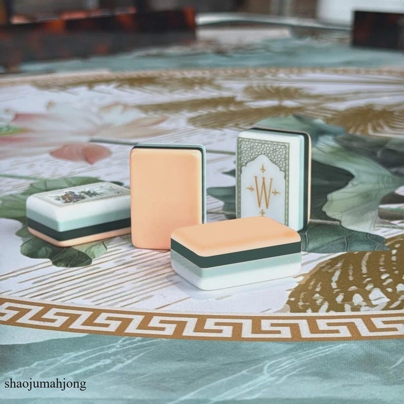 May include: Four rectangular mahjong tiles featuring a layered design with a peach-colored top, a dark green middle, and a light green base. One tile displays a white face with a gold letter W and decorative green border, while another shows a floral design.
