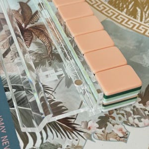 May include: A clear acrylic rack holding a stack of rectangular tiles. The tiles feature a smooth peach-colored top surface with layered green and white edges. The transparent rack has a visible logo etched into the center of its base surface.