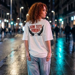 Op de afbeelding: Een wit t-shirt met de tekst "SOM TOTS DIMONIS" in rood en zwart, met een stierenkopontwerp. Het shirt wordt gedragen met lichtblauwe jeans. De foto is 's nachts buiten genomen, met straatverlichting op de achtergrond.