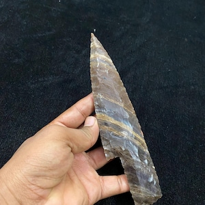 以下が含まれることがあります： 手持ちの茶色と灰色の矢じりで、鋭い先端と切り欠きのある基部があります。矢じりは層状のストライプ模様で、石のように見えます。背景は暗く、質感のある表面です。