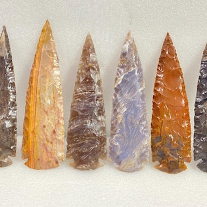May include: Six polished stone arrowheads in shades of brown, orange, and purple are arranged in a row. Each arrowhead has a triangular shape with a pointed tip and a notched base. The stones have a glossy finish.