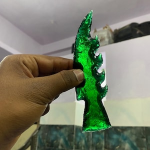 Puede incluir: Una escultura de vidrio abstracta, translúcida, de color verde esmeralda, con una forma orgánica y dentada. El objeto se sostiene sobre un fondo neutro, mostrando su superficie texturizada y sus cualidades de captura de la luz. La pieza tiene un diseño artístico único.