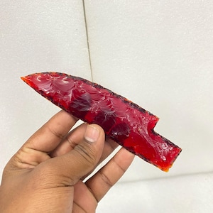 May include: A translucent, red arrowhead with a textured surface. The arrowhead has a sharp point and a notched base. The color is a deep, rich red, with hints of orange at the edges. The arrowhead is held in a hand against a white background.