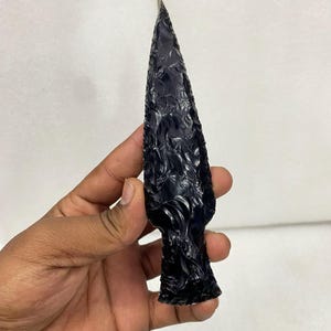 Puede incluir: Una punta de flecha de obsidiana negra con una punta afilada y superficie texturizada. La punta de flecha es de un negro oscuro y brillante, y parece estar tallada a mano. Es un artefacto histórico.