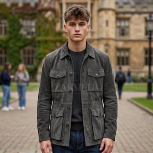 Puede incluir: Chaqueta de ante gris con cuatro bolsillos delanteros y cierre de botones. La chaqueta se lleva sobre una camiseta de cuello redondo gris oscuro y vaqueros azules. La chaqueta tiene un diseño clásico y a medida.