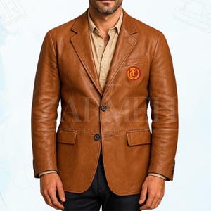 May include: A brown leather blazer with a classic two-button closure and notched lapels. The blazer features a small emblem on the chest and two front pockets. The jacket is worn over a tan button-down shirt.