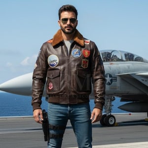 May include: A brown leather bomber jacket with a tan faux fur collar, featuring multiple colorful embroidered patches on the chest and sleeves, a front zipper closure, and ribbed cuffs, worn with blue denim jeans and a black leg holster.