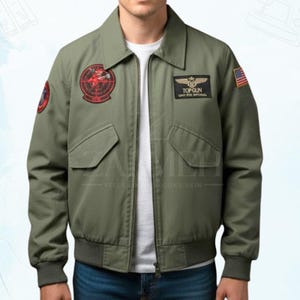 Könnte beinhalten: Eine olivgrüne Bomberjacke mit durchgehendem Frontreißverschluss, spitzem Kragen sowie gerippten Bündchen und Saum. Die Jacke verfügt über zwei Brusttaschen mit Klappen, einen Top Gun-Aufnäher, einen runden rot-schwarzen Aufnäher und eine US-Flagge.