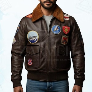 Könnte beinhalten: Braune Leder-Bomberjacke mit braunem Kunstfellkragen und mehreren gestickten Aufnähern. Die Jacke hat zwei Vordertaschen und einen Reißverschluss. Der Text "Armeh" und "Feels like a second skin" sind sichtbar.