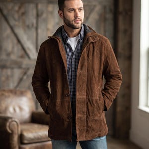 May include: A brown suede jacket featuring a high collar, a front button closure, and two side pockets. The jacket is worn over a white t-shirt and a blue and white checkered button-down shirt, paired with blue denim jeans.