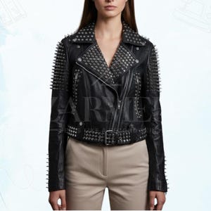 May include: Black leather jacket with silver stud and spike embellishments. The jacket features a notched collar, asymmetrical zipper closure, and a studded belt. The shoulders and collar are heavily studded, and the sleeves have spike accents. The jacket is paired with tan pants.