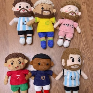 May include: Six crocheted dolls representing soccer players in various team uniforms. The dolls feature embroidered facial details, beards, and jerseys with numbers like 7 and 10. The uniforms include light blue and white stripes, yellow, pink, red, and dark blue colors.